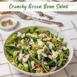 A white bowl holds a vibrant green bean salad with lettuce, red onion, crumbled white cheese, and sliced almonds, on a checkered napkin with silverware.