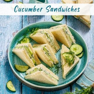 A teal plate holds several triangular cucumber sandwiches garnished with dill and cucumber slices, on a blue distressed wooden table.