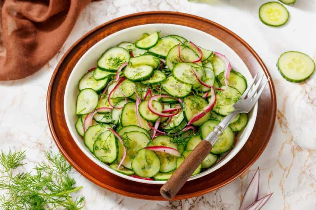 A bowl of cucumber salad with red onions and fresh dill, placed on a wooden plate with a fork, surrounded by ingredients on a marble surface.