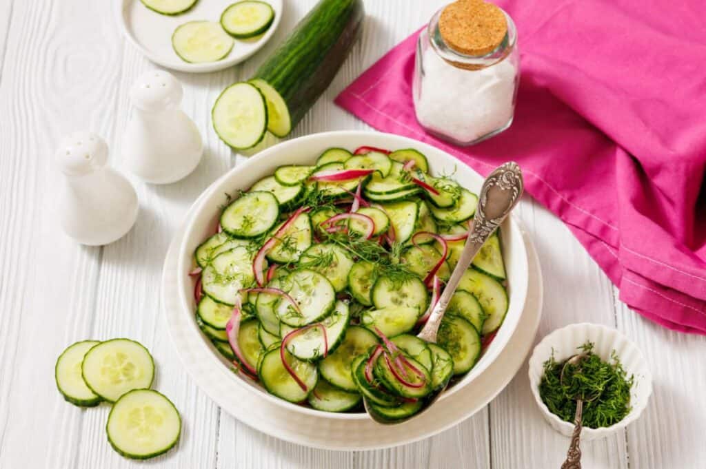 A white bowl filled with cucumber and red onion salad garnished with dill, placed on a white table with a pink napkin, salt, pepper shakers, and fresh cucumber slices nearby.
