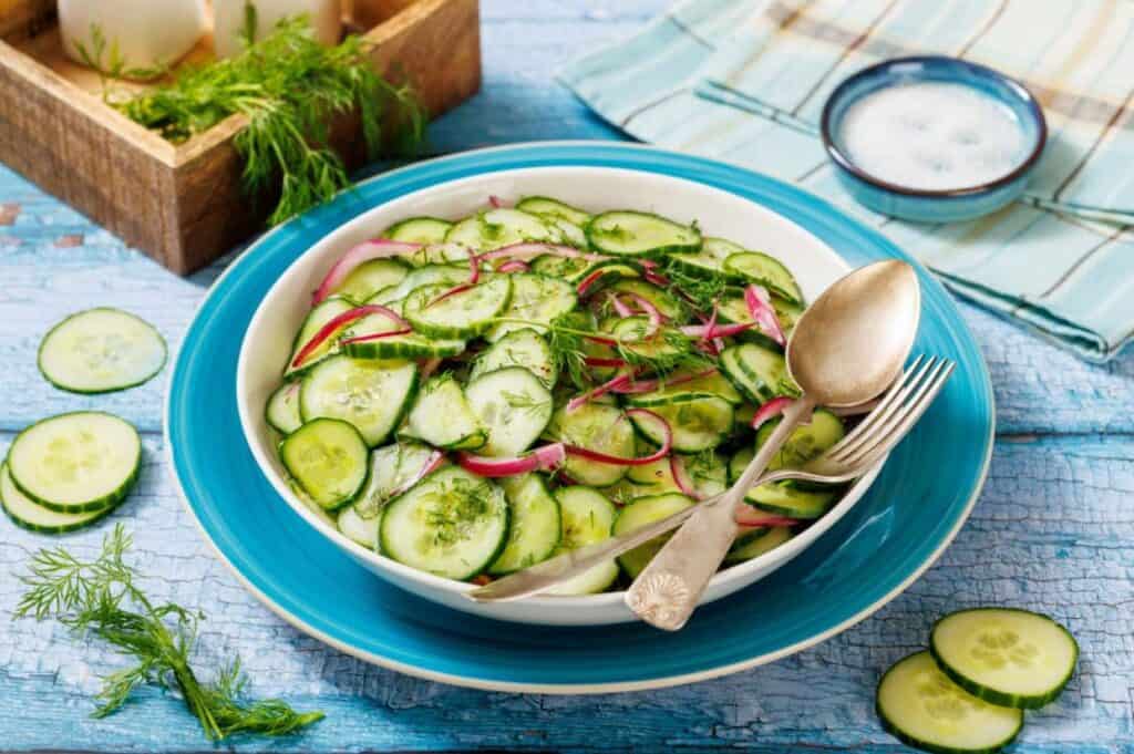 A bowl of cucumber and red onion salad garnished with dill, placed on a blue plate with a fork and spoon, on a blue wooden surface.