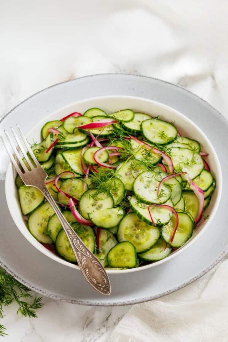 A white bowl filled with sliced cucumbers, red onions, and fresh dill sits on a plate with a fork resting on the edge.