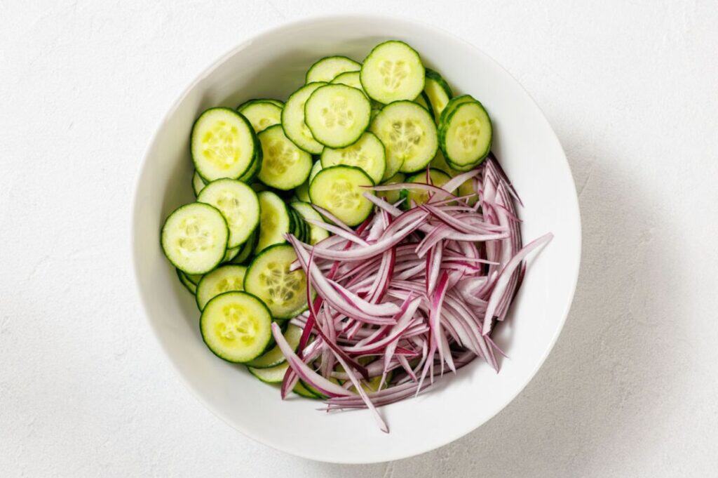 A white bowl containing sliced cucumbers and thinly sliced red onions on a white surface.