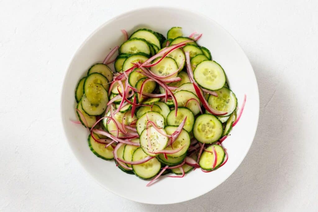 A white bowl filled with sliced cucumbers and thinly sliced red onions, seasoned with black pepper.