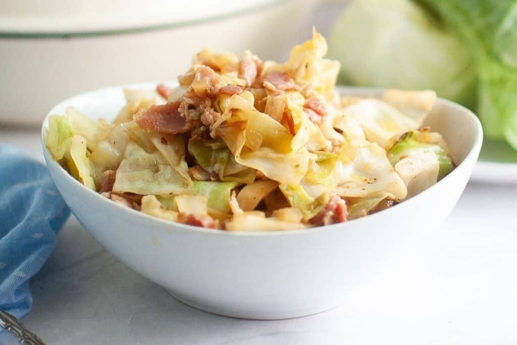 A white bowl brimming with golden-brown fried cabbage and crispy bits of bacon, with a blue napkin and a large white pot in the blurred background.