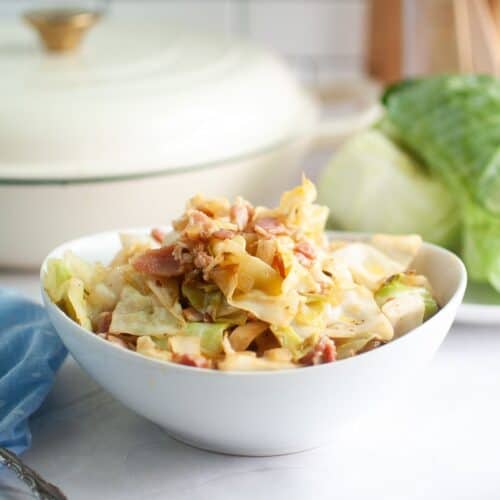 A white bowl brimming with golden-brown fried cabbage and crispy bits of bacon, with a blue napkin and a large white pot in the background.