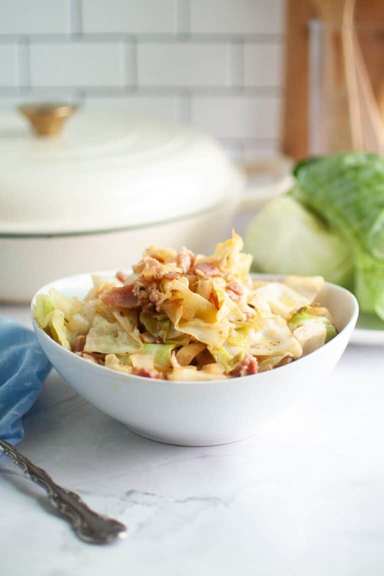 A white bowl brimming with golden-brown fried cabbage and crispy bits of bacon, with a blue napkin and a large white pot in the background.