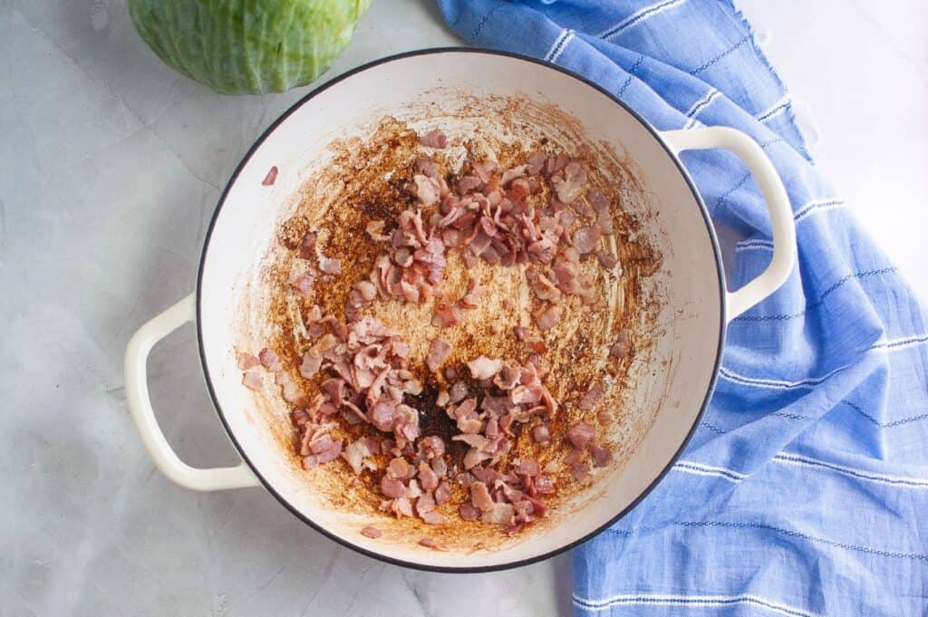 An overhead view of a white enameled cast iron pan containing crispy, browned bits of cooked bacon and rendered fat, with a blue and white striped napkin and a head of green cabbage in the background.