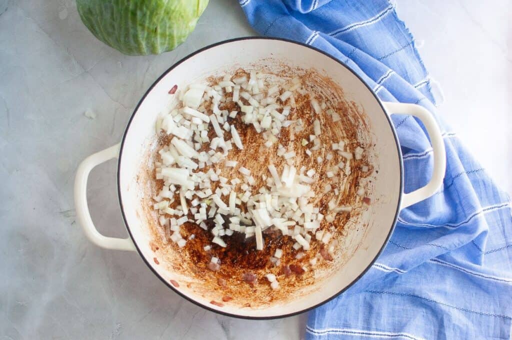 An overhead view of a white enameled pan with crispy cooked bacon bits and rendered fat, now with a layer of freshly diced white onions scattered over them, with a blue and white striped napkin and green cabbage in the background.