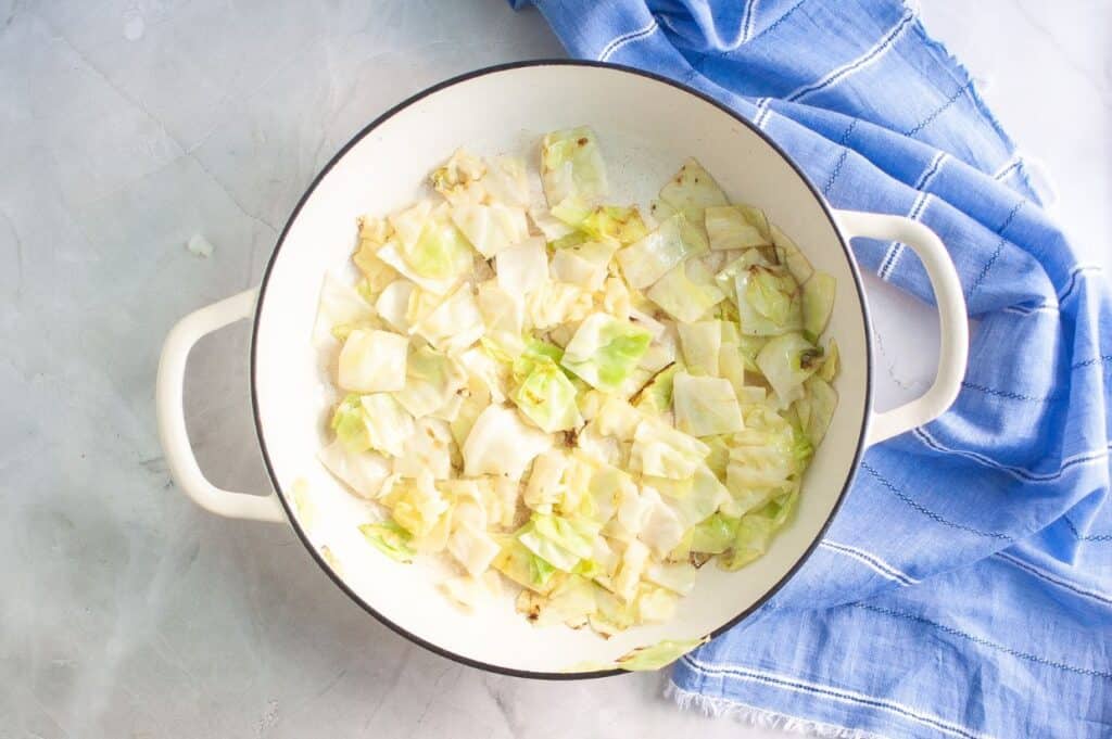An overhead view of a white enameled pan filled with a generous amount of chopped, tender, light green and golden-brown cooked cabbage, with a blue and white striped napkin in the background.