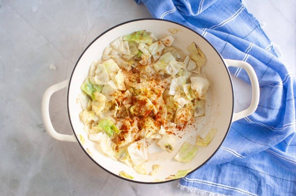 An overhead view of a white enameled pan containing tender, light green and golden-brown cooked cabbage pieces, generously sprinkled with reddish-orange paprika, with a blue and white striped napkin in the background.