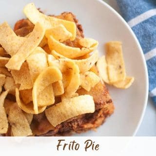 A serving of Frito pie topped with corn chips on a white plate, next to a blue and white striped cloth.