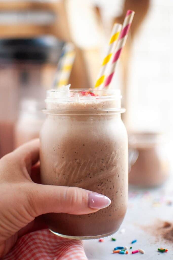 A hand holds a clear mason jar filled with frothy brown frozen hot chocolate, topped with whipped cream, red sprinkles, and two colorful striped straws.
