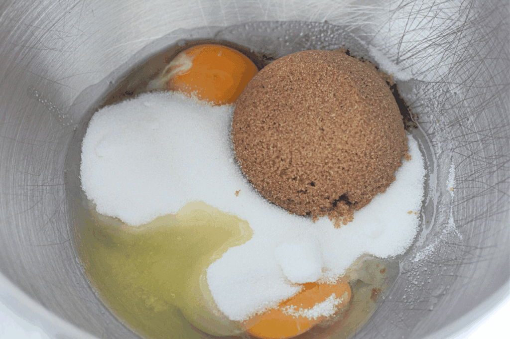 A metal mixing bowl containing eggs, granulated white sugar, and a mound of brown sugar.