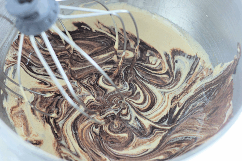 A close-up of a stand mixer whisk attachment mixing chocolate and vanilla batter in a metal bowl, creating a marbled swirl pattern.