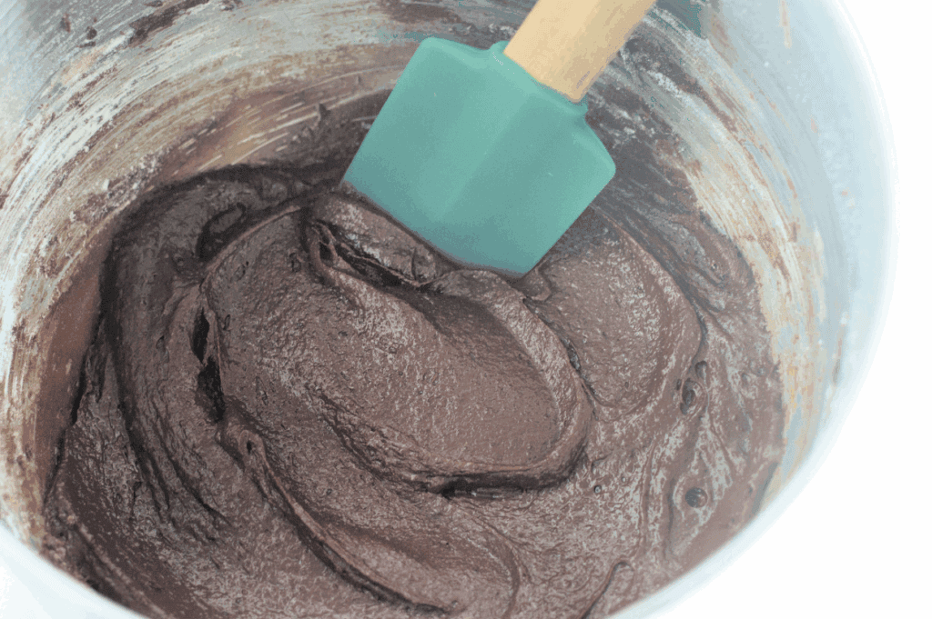 A green spatula mixing thick chocolate batter in a metal bowl.