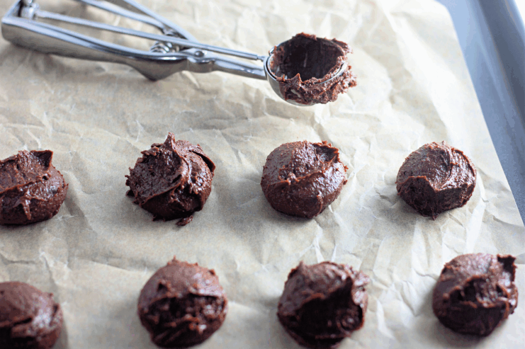 Spoonfuls of chocolate cookie dough are arranged on a parchment-lined baking sheet, with a metal scoop resting nearby.