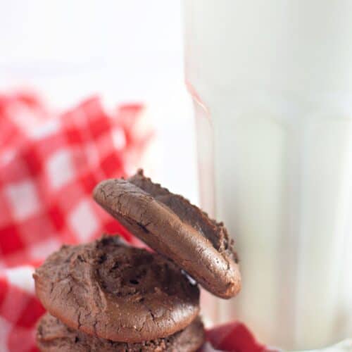 A stack of chocolate cookies on a red and white checkered cloth next to a glass of milk.
