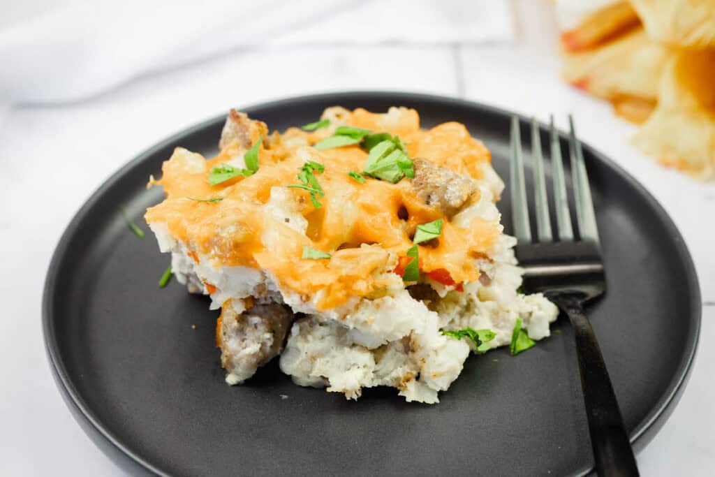 A serving of cheesy breakfast casserole with sausage and chopped herbs on a black plate, next to a black fork.