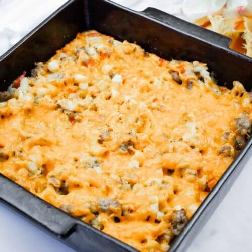 A black square baking dish filled with baked macaroni casserole topped with melted cheese sits on a white surface next to a white and yellow floral arrangement.