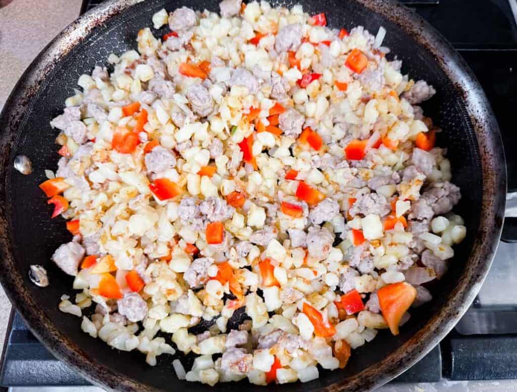 A frying pan filled with chopped onions, red bell peppers, cauliflower, and sausage pieces cooking on a stovetop.