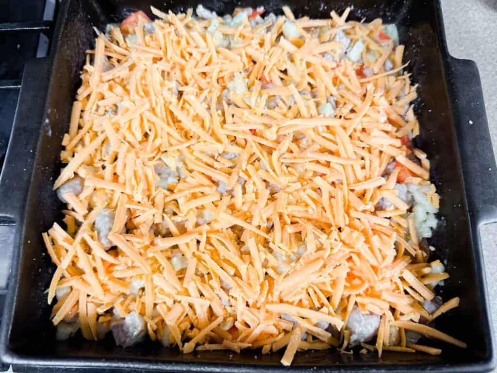 A square baking dish filled with a mixture of chopped vegetables and meat, topped with a generous layer of shredded cheddar cheese.