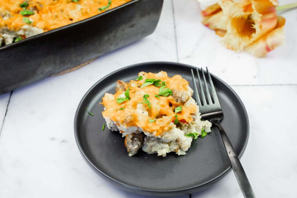 A serving of casserole with cheese, ground meat, and herbs on a black plate with a fork, next to a casserole dish and pasta shells on a marble surface.