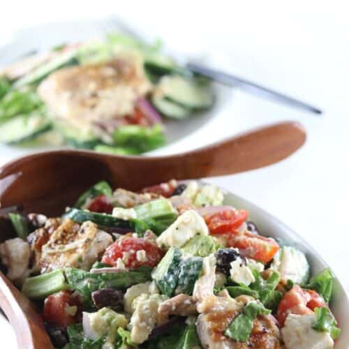 A bowl of Greek salad with grilled chicken, tomatoes, cucumbers, olives, feta cheese, and greens, served with wooden salad servers. A plate with salad is in the background.