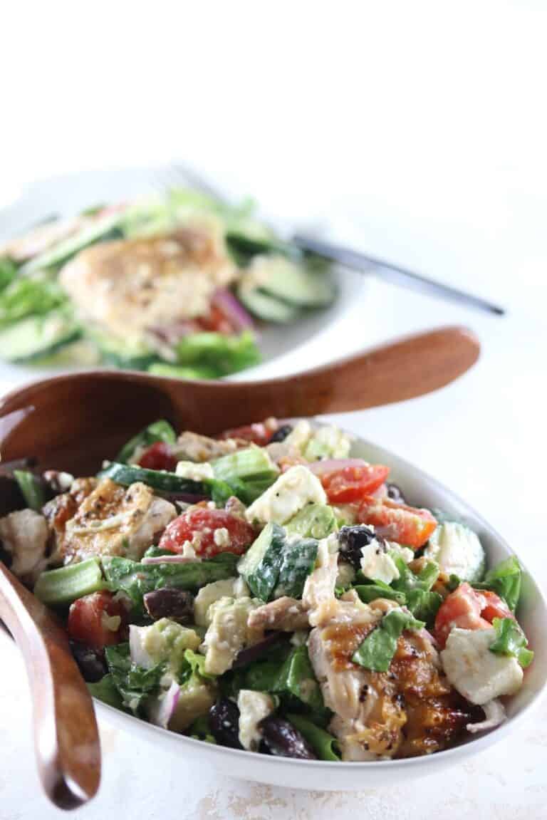 A bowl of Greek salad with grilled chicken, tomatoes, cucumbers, olives, feta cheese, and greens, served with wooden salad servers. A plate with salad is in the background.
