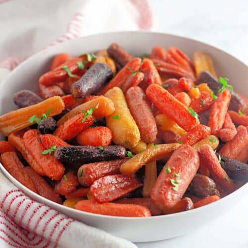 A white bowl filled with assorted roasted baby carrots garnished with chopped herbs, placed on a white surface with a red-striped cloth nearby.