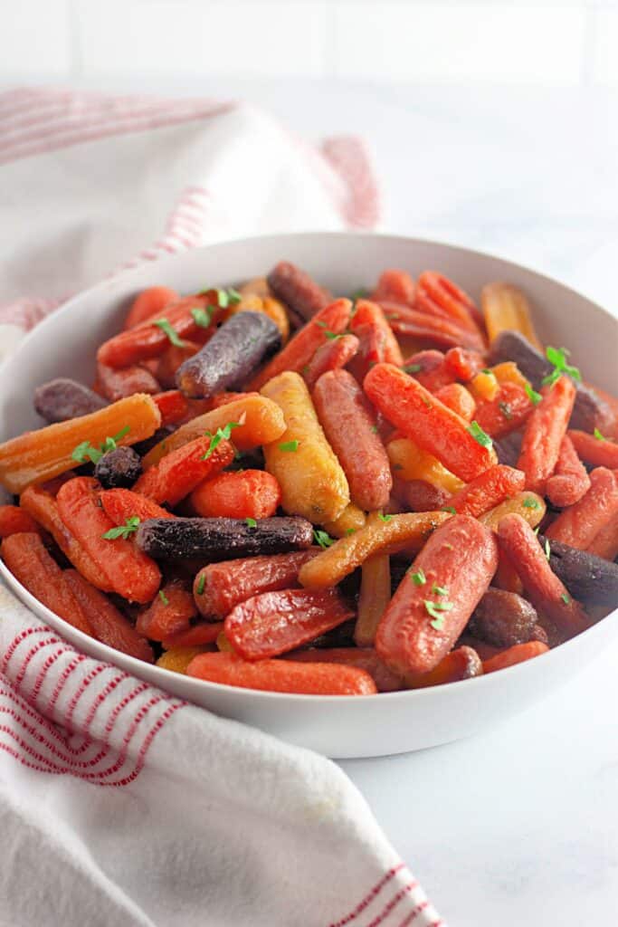 A white bowl filled with assorted roasted baby carrots garnished with chopped herbs, placed on a white surface with a red-striped cloth nearby.