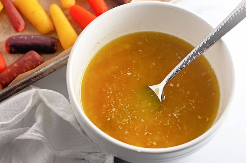 A bowl of yellow sauce or dressing with a spoon, placed on a white surface next to a tray of colorful carrots and a white cloth.