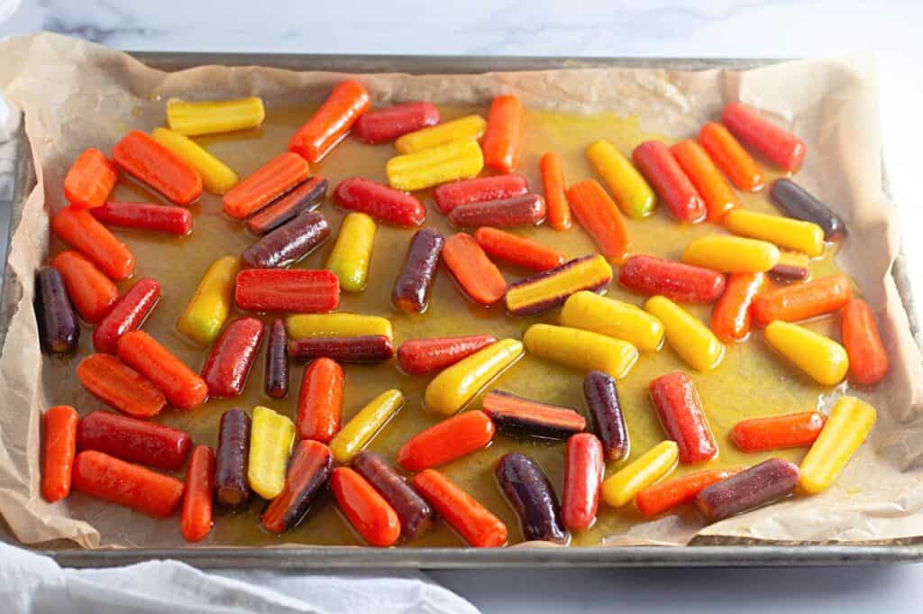 A baking sheet lined with parchment paper holds colorful orange, yellow, red, and purple carrots coated with oil, ready for roasting.