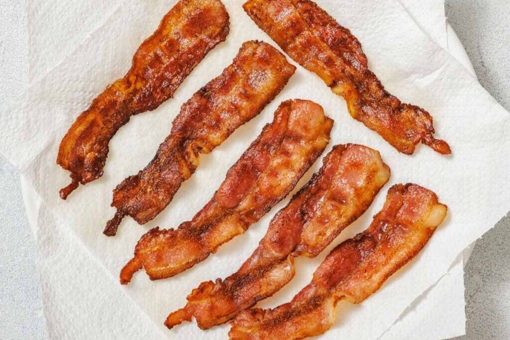 Six strips of cooked bacon are laid out on a white paper towel to absorb excess grease.