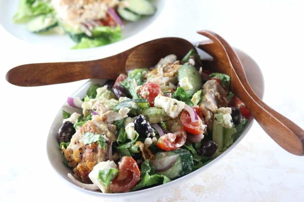 A bowl of Greek salad with grilled chicken, cherry tomatoes, cucumbers, olives, red onions, feta cheese, and wooden salad servers.