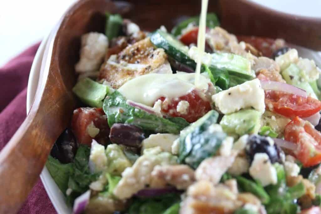 A close-up of a Greek salad with lettuce, tomatoes, cucumbers, olives, red onions, grilled chicken, and feta cheese, topped with a creamy dressing being poured.