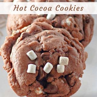 A close-up of soft, dark brown hot cocoa cookies, studded with chocolate chips and topped with small white marshmallows, on a light-colored surface.