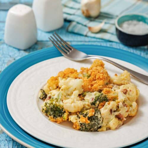 A plate of baked broccoli and cauliflower casserole sits on a blue and white plate with a fork, on a rustic blue table beside salt, pepper, and a napkin.