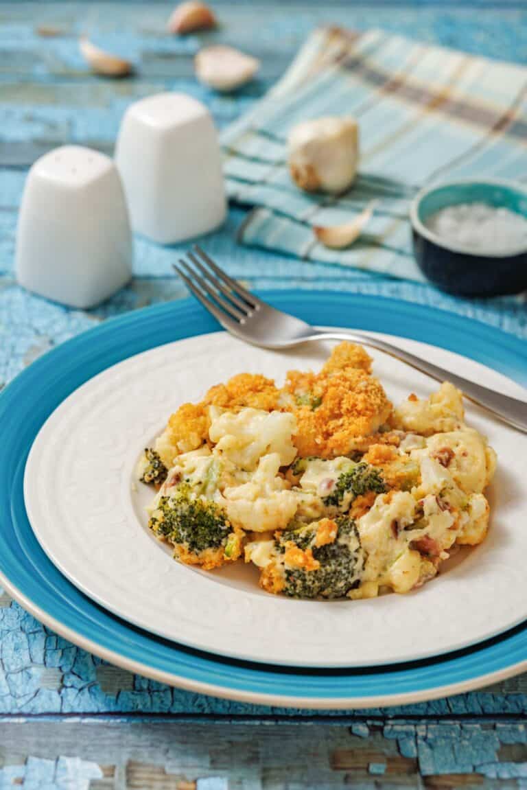A plate of baked broccoli and cauliflower casserole sits on a blue and white plate with a fork, on a rustic blue table beside salt, pepper, and a napkin.