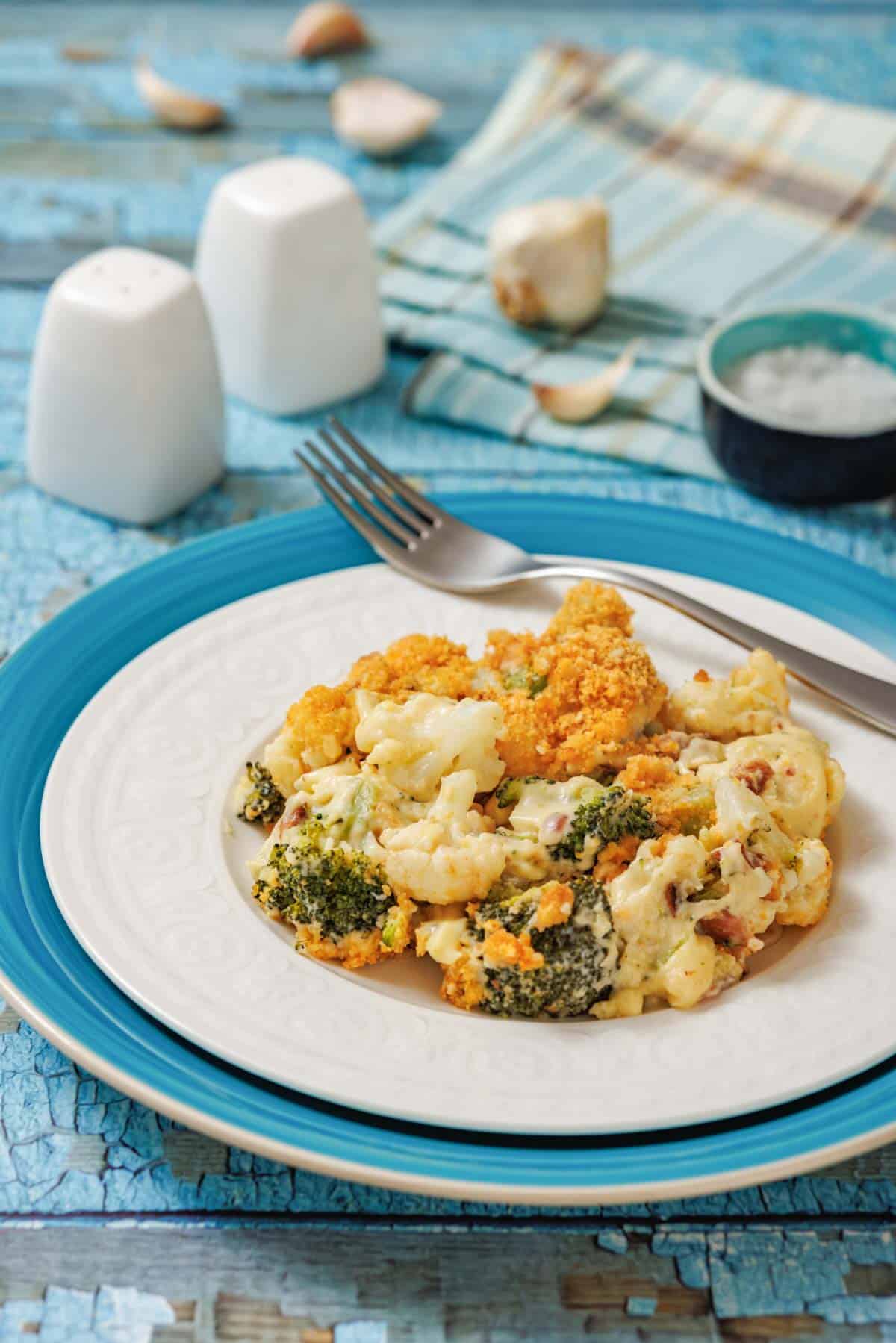 A plate of baked broccoli and cauliflower casserole sits on a blue and white plate with a fork, on a rustic blue table beside salt, pepper, and a napkin.