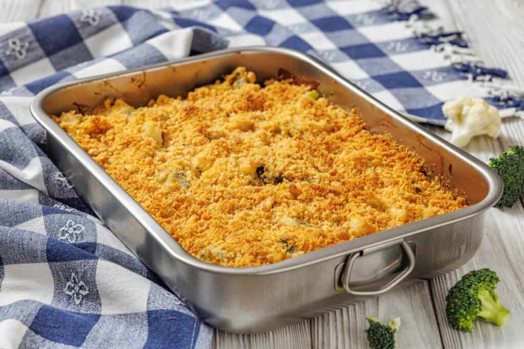 A rectangular baking dish filled with a golden-brown breadcrumb-topped casserole sits on a blue and white checkered cloth, with broccoli and cauliflower pieces nearby.