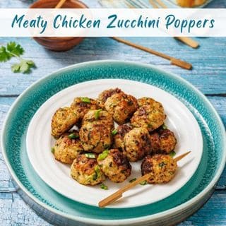 A white plate on a teal charger holds several golden-brown Meaty Chicken Zucchini Poppers garnished with green onions, with wooden skewers on a blue distressed wooden table.