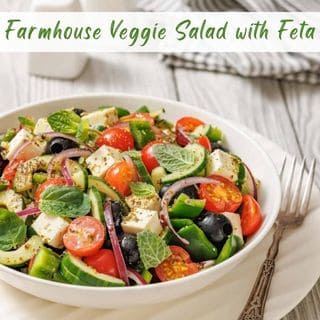 A white bowl is filled with a colorful mix of chopped vegetables, olives, and feta cheese, dressed with herbs, with a fork on the side.