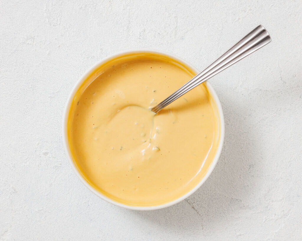 A bowl filled with creamy yellow cheese sauce, with a metal spoon resting inside, on a light textured surface.
