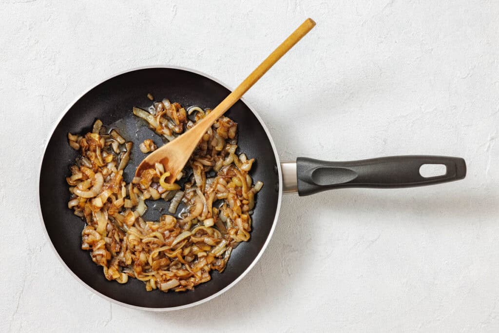 Sautéed onions in a black frying pan, stirred with a wooden spoon, on a white textured surface.