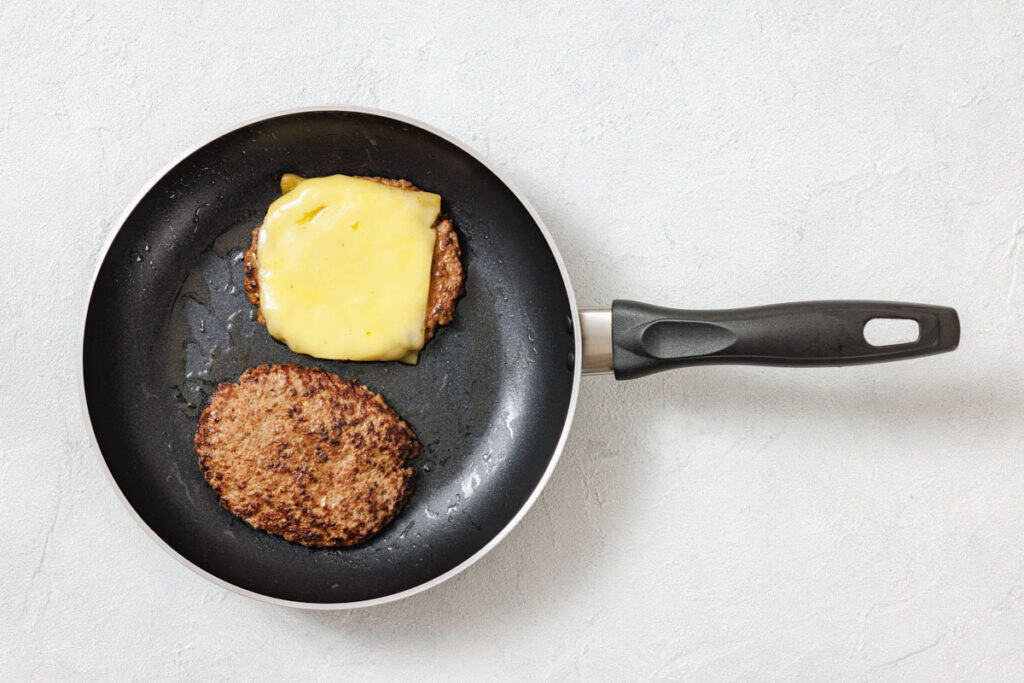 Two cooked burger patties in a frying pan, one topped with melted cheese, on a light-colored surface.