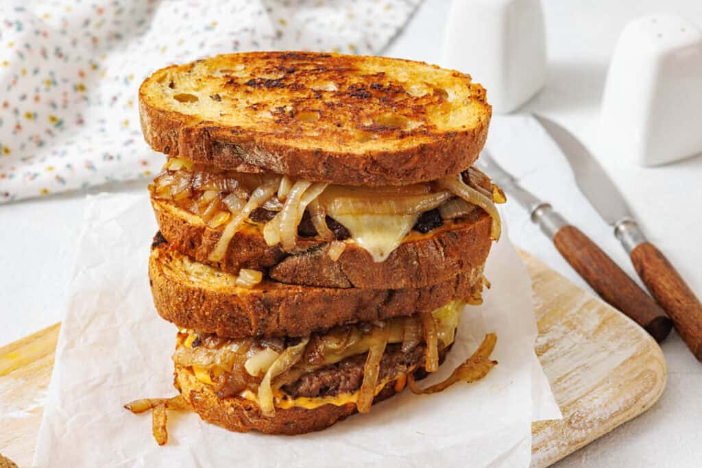 A stack of two grilled cheese sandwiches with caramelized onions and melted cheese on toasted bread, placed on parchment paper atop a wooden board.