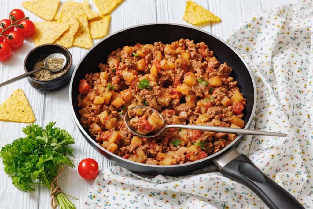 Overhead shot of a black skillet filled with seasoned ground meat and diced potatoes, garnished with parsley, alongside tortilla chips and tomatoes.