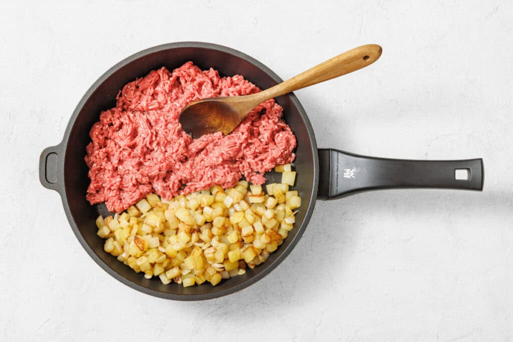 Overhead shot of a black nonstick skillet divided, with raw ground meat on one side and golden-brown diced potatoes on the other, with a wooden spoon in the meat.
