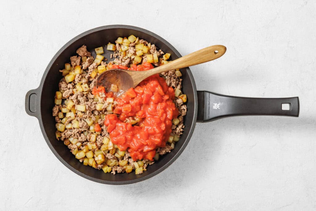 Overhead shot of a black nonstick skillet filled with cooked ground meat and diced potatoes, with a pile of diced tomatoes in the center and a wooden spoon.